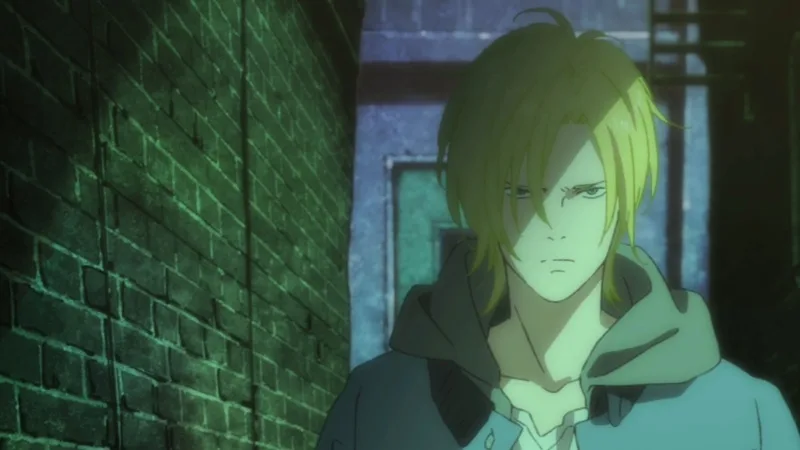 The image features a close-up of the character Ash Lynx, the protagonist of the anime series Banana Fish. He has short, messy, light-blond or yellow hair and intense green eyes. He is wearing a light blue or grey hoodie over a white collared shirt. The background is dark, appearing to be an alleyway or a dimly lit outdoor area. A prominent green brick wall is on the left, and a dark blue or grey wall with a door is on the right.