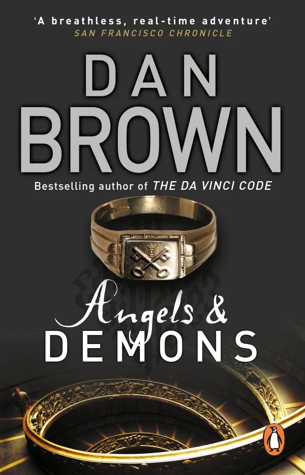 Description: The cover of the book Angels & Demons by Dan Brown. Text content: The cover features the author's name, the book title, and the tagline "Bestselling author of THE DA VINCI CODE". Visuals: A prominent gold ring featuring a symbol of crossed keys and a papal tiara is centered on the cover.