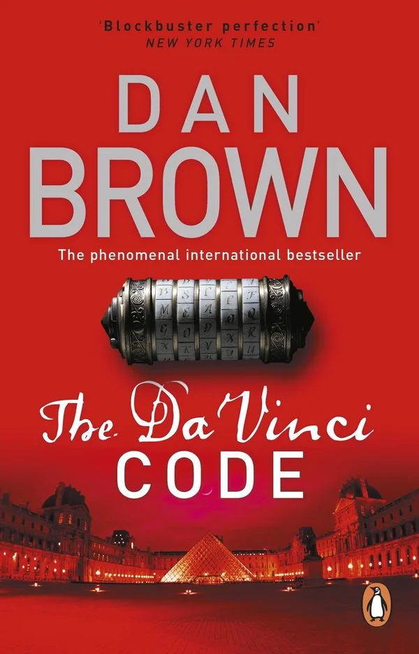 The image is the cover of the internationally bestselling novel The Da Vinci Code. Visuals: The background is bright red. The author's name, "DAN BROWN," is prominent at the top in large white letters. The title, "The Da Vinci CODE," is displayed below in a stylized font. A metallic cryptex, a portable vault used to hide secret messages in the story, is featured in the center of the cover. Setting: The bottom of the cover shows an illustration of the Louvre museum and its pyramid in Paris, which is a key location in the book's plot. Text: Textual elements include the tagline "'Blockbuster perfection' NEW YORK TIMES" and "The phenomenal international bestseller". 