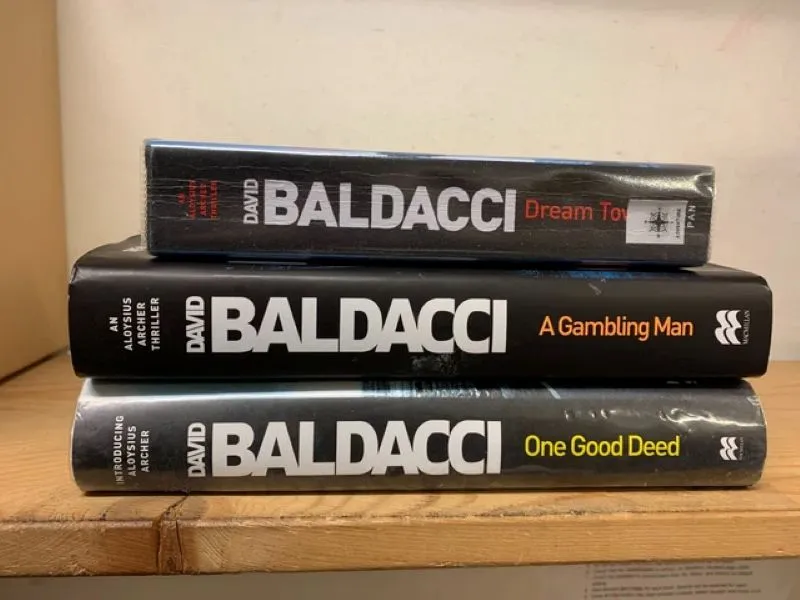 Three hardcover books by David Baldacci stacked horizontally on a wooden surface: 'Dream Town', 'A Gambling Man', and 'One Good Deed'.