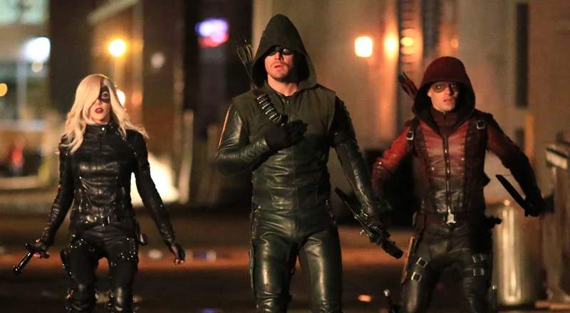 An image of three people in superhero costumes standing on a street at night. The central figure is Oliver Queen as the Green Arrow, flanked by Black Canary and Arsenal. All are wearing their full vigilante gear, including masks and weapons. 
