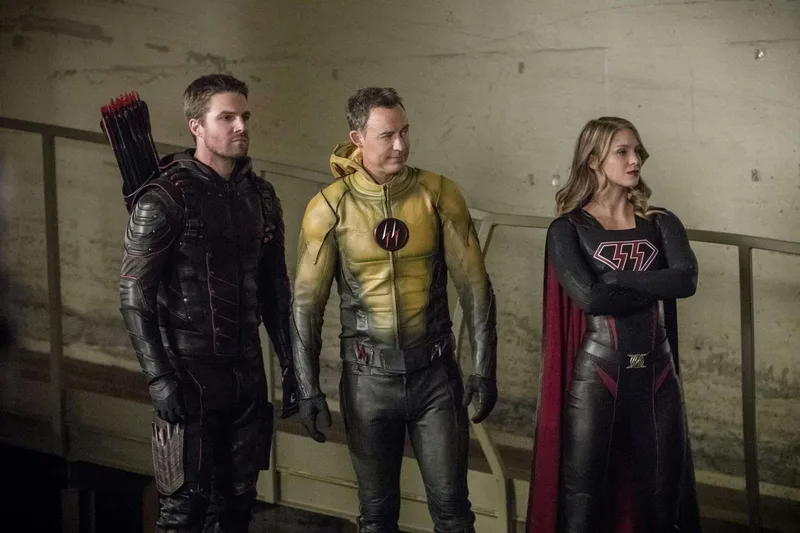 Three characters from the "Crisis on Earth-X" crossover stand together in a concrete area. On the left is Earth-X's Dark Arrow (Oliver Queen doppelganger) in a black leather suit with a quiver of arrows on his back. In the center is Earth-X's Reverse-Flash (Eobard Thawne as Harrison Wells doppelganger) in a yellow and black suit. On the right is Earth-X's Overgirl (Kara Zor-El doppelganger) in a black and red Supergirl-style costume with a red cape. 