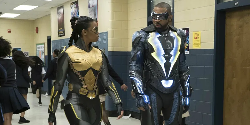 Description: The image shows two individuals dressed in superhero costumes standing in a school hallway. Characters: The man on the right is Jefferson Pierce (Black Lightning), wearing a black and blue suit with a mask. The woman on the left is Anissa Pierce, wearing a black and yellow suit. Setting: They are in a school hallway, with a sign reading "PRIDE" visible on the wall in the background.