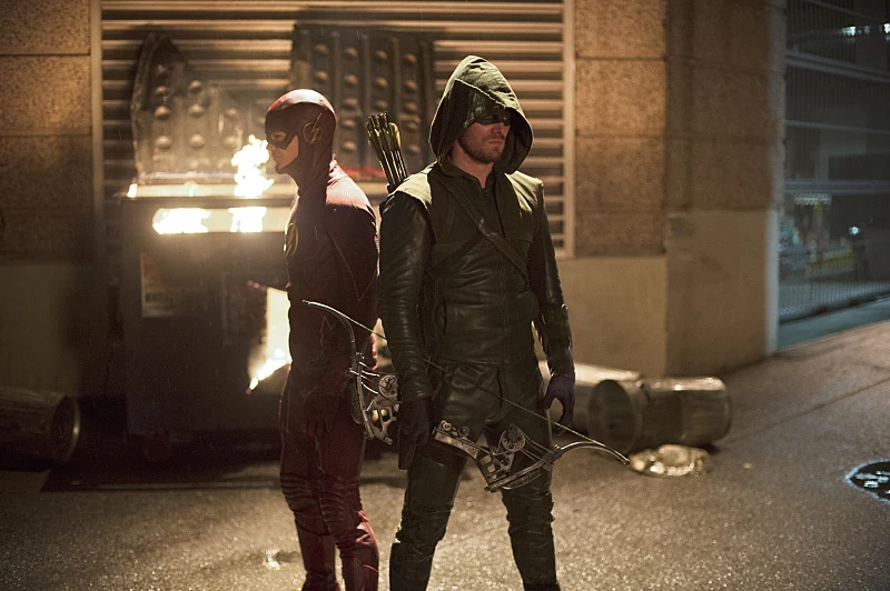 This picture displays the characters The Flash and Green Arrow standing back-to-back in an alleyway at night. The character on the left is The Flash, wearing a red suit, while the character on the right is Green Arrow, wearing a green hooded leather suit and holding a bow.