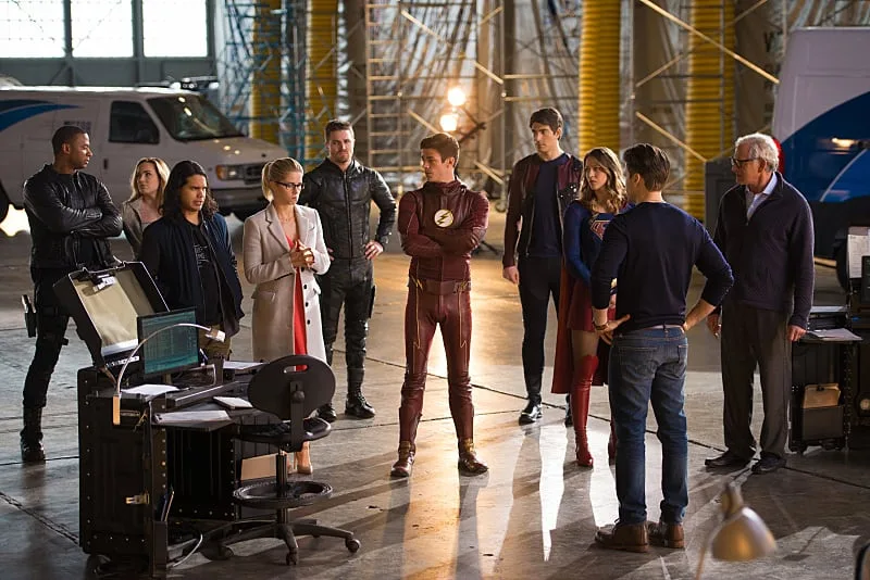 A group of characters from the CW superhero shows, including The Flash (Grant Gustin) in his red suit, Supergirl (Melissa Benoist) in her blue and red suit, and Green Arrow (Stephen Amell) in civilian clothes, stand together in a large, industrial warehouse setting with other cast members.