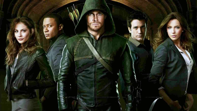 A promotional image for the TV series Arrow showing five main characters. In the center stands the Green Arrow (Oliver Queen) in his signature green hooded leather costume, holding a bow and with a quiver of arrows visible over his shoulder. To his left (viewer's right) is a woman in a black jacket. To his right (viewer's left) are three other characters: a woman in a black leather jacket, a man in a black suit jacket, and another man in a grey suit jacket. The background is dark and indistinct. 