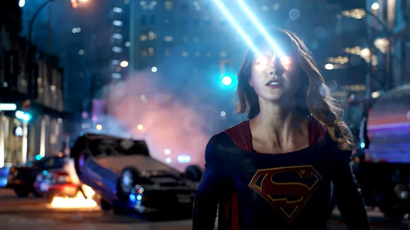 A still from the Supergirl TV series shows the character Kara Zor-El (played by Melissa Benoist) using her heat vision. The image captures her in her Supergirl costume, with bright red and orange light emanating from her eyes as blue beams shoot upward. The setting is a night-time city street, with a flipped car visible in the background and a blue light in the mid-ground. The scene is dramatic and action-oriented, likely from an episode such as "Hostile Takeover". 