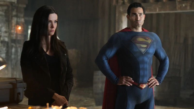 A man in a blue and red Superman suit with a red cape stands next to a woman with long dark hair wearing a black jacket, likely actors Tyler Hoechlin and Elizabeth Tulloch from the series Superman & Lois. They are standing in what appears to be a barn or rustic indoor setting. 