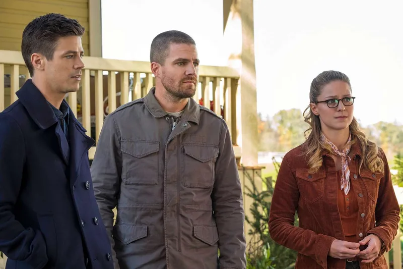 Description: Grant Gustin (left), Stephen Amell (center), and Melissa Benoist (right) stand outdoors during the "Elseworlds" Arrowverse crossover event. The characters appear to be on a porch or deck. Context: This image is from The Flash Season 5, Episode 9, titled "Elseworlds, Part 1", where the characters Barry Allen and Oliver Queen have swapped bodies. Grant Gustin is playing Oliver Queen's character, and Stephen Amell is playing Barry Allen's character. Melissa Benoist is playing Kara Danvers/Supergirl.